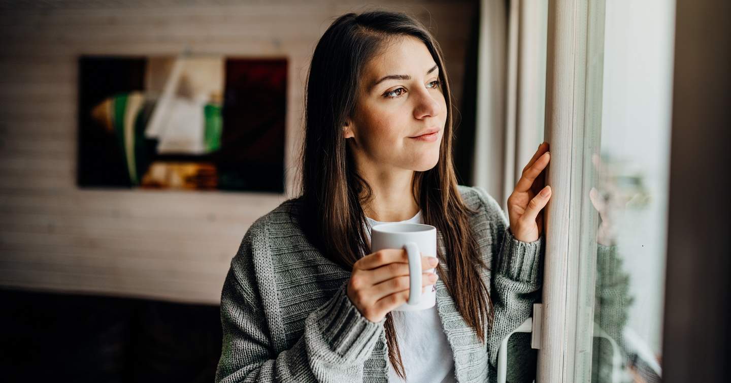 person looking forlorn out the window with a cup of a hot liquid in their hands